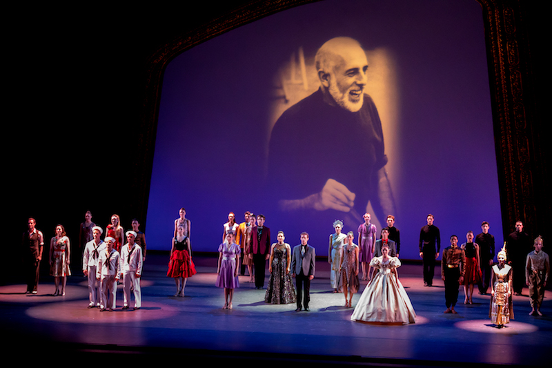Dancers stand in costumes, including sailor suits and evening gowns, in front of a video projection that features an image of the late Jerome Robbins